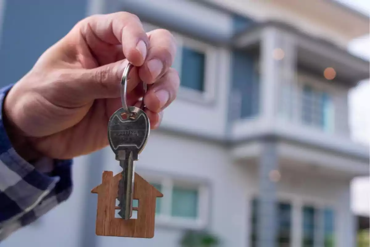 Person holding a key with a house-shaped keychain in front of a blurred modern home