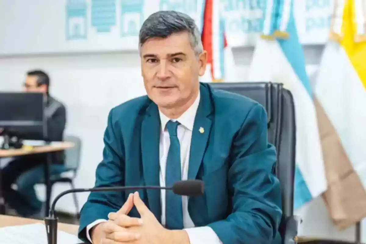 Hombre de cabello canoso con traje azul sentado en una oficina frente a un micrófono con banderas de fondo. Hombre de cabello canoso con traje azul sentado en una oficina frente a un micrófono con banderas de fondo.