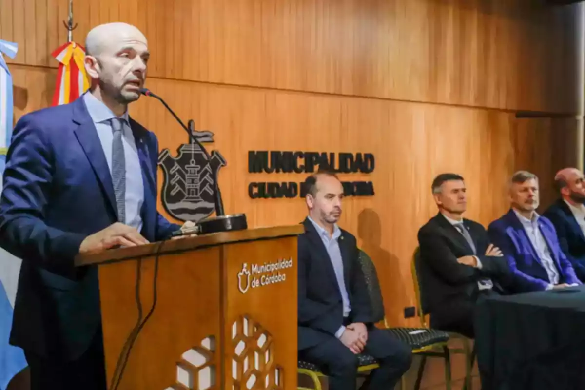 A man in a suit speaks at a podium during an official event at the Municipality of Córdoba while four other people in formal attire are seated beside him in a room with wooden walls. A man in a suit speaks at a podium during an official event at the Municipality of Córdoba while four other people in formal attire are seated beside him in a room with wooden walls.