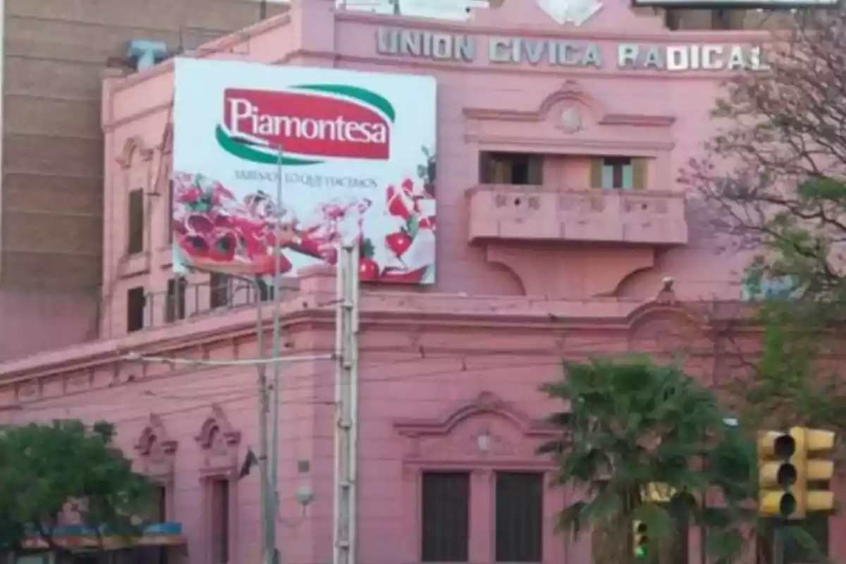 Edificio rosa de la Unión Cívica Radical con un cartel publicitario de Piamontesa en la fachada y un semáforo en primer plano Edificio rosa de la Unión Cívica Radical con un cartel publicitario de Piamontesa en la fachada y un semáforo en primer plano