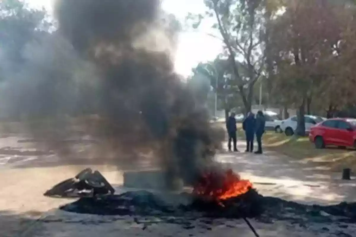 Humo negro y fuego saliendo de una pila de neumáticos quemados en una calle con algunos autos y personas al fondo Humo negro y fuego saliendo de una pila de neumáticos quemados en una calle con algunos autos y personas al fondo