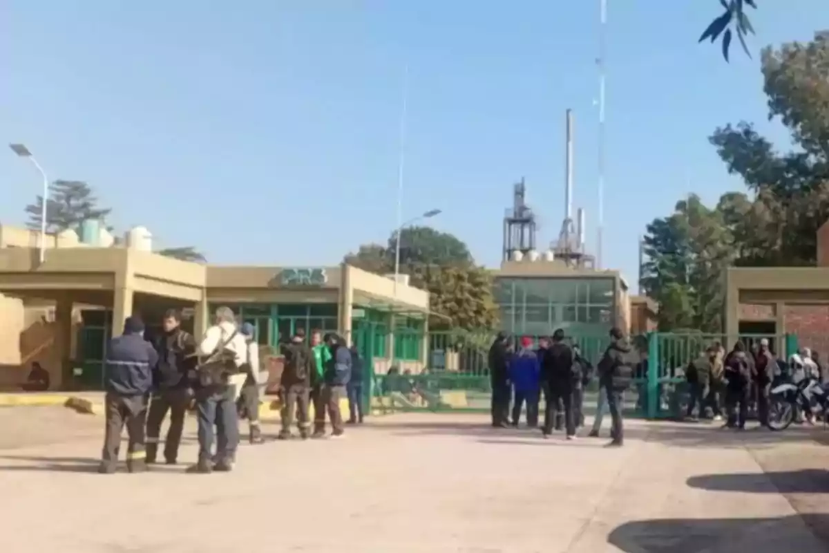 Varias personas se encuentran reunidas frente a la entrada de una planta industrial con portones verdes y edificios de fondo en un día soleado. Varias personas se encuentran reunidas frente a la entrada de una planta industrial con portones verdes y edificios de fondo en un día soleado.
