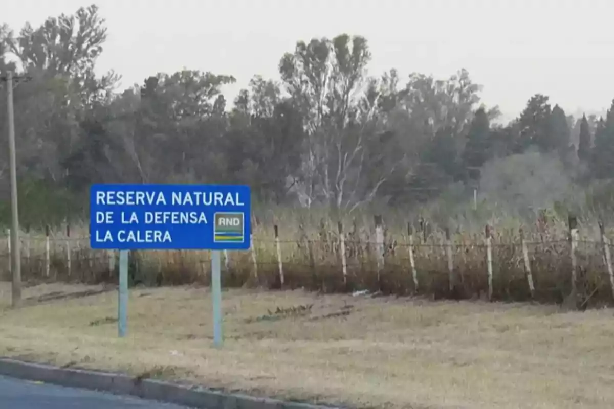 Cartel azul que indica la entrada a la Reserva Natural de la Defensa La Calera junto a un camino y vegetación de fondo Cartel azul que indica la entrada a la Reserva Natural de la Defensa La Calera junto a un camino y vegetación de fondo