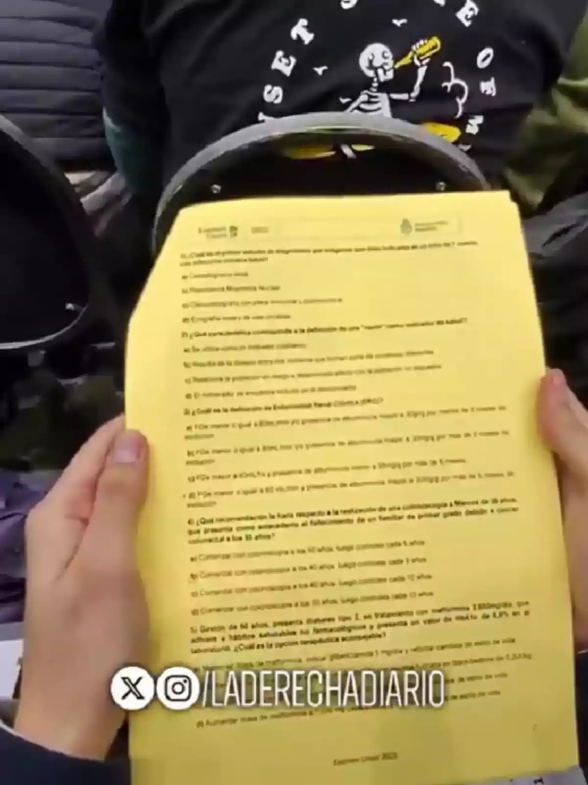 Persona sosteniendo una hoja amarilla con preguntas impresas en un aula, al fondo se ve a otra persona con una camiseta negra con un dibujo de esqueleto Persona sosteniendo una hoja amarilla con preguntas impresas en un aula, al fondo se ve a otra persona con una camiseta negra con un dibujo de esqueleto