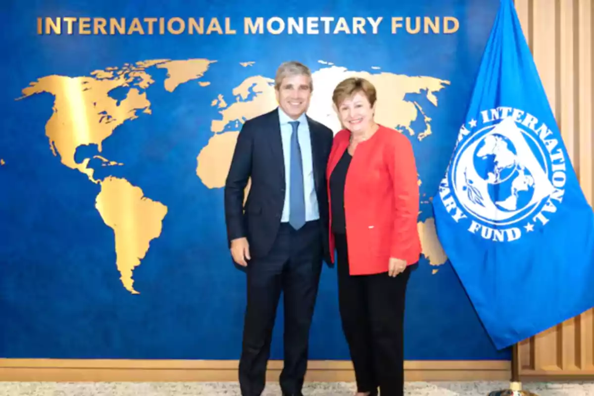 Dos personas posan sonrientes frente a un fondo azul con un mapa dorado del mundo y el texto International Monetary Fund junto a una bandera azul del mismo organismo