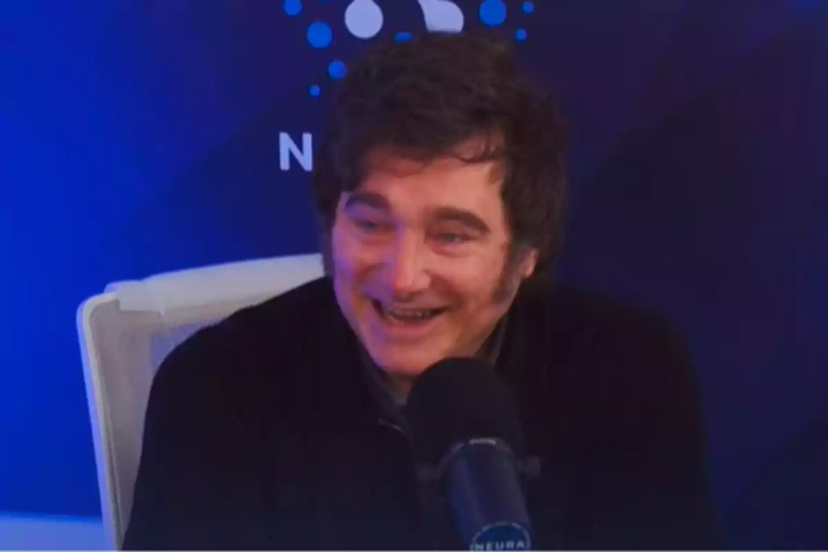 Hombre de cabello oscuro sonriendo mientras habla frente a un micrófono en un estudio con fondo azul