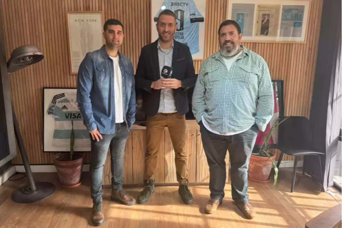 Tres hombres posan en una oficina decorada con camisetas deportivas enmarcadas, plantas y recortes de periódico en la pared