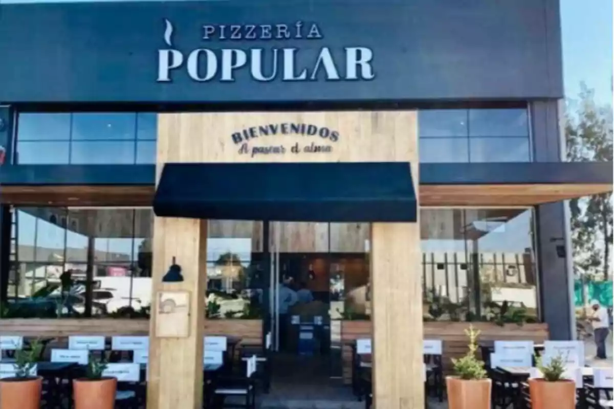 Fachada de una pizzería llamada Popular con mesas y sillas en el exterior y un letrero de bienvenida sobre la entrada Fachada de una pizzería llamada Popular con mesas y sillas en el exterior y un letrero de bienvenida sobre la entrada