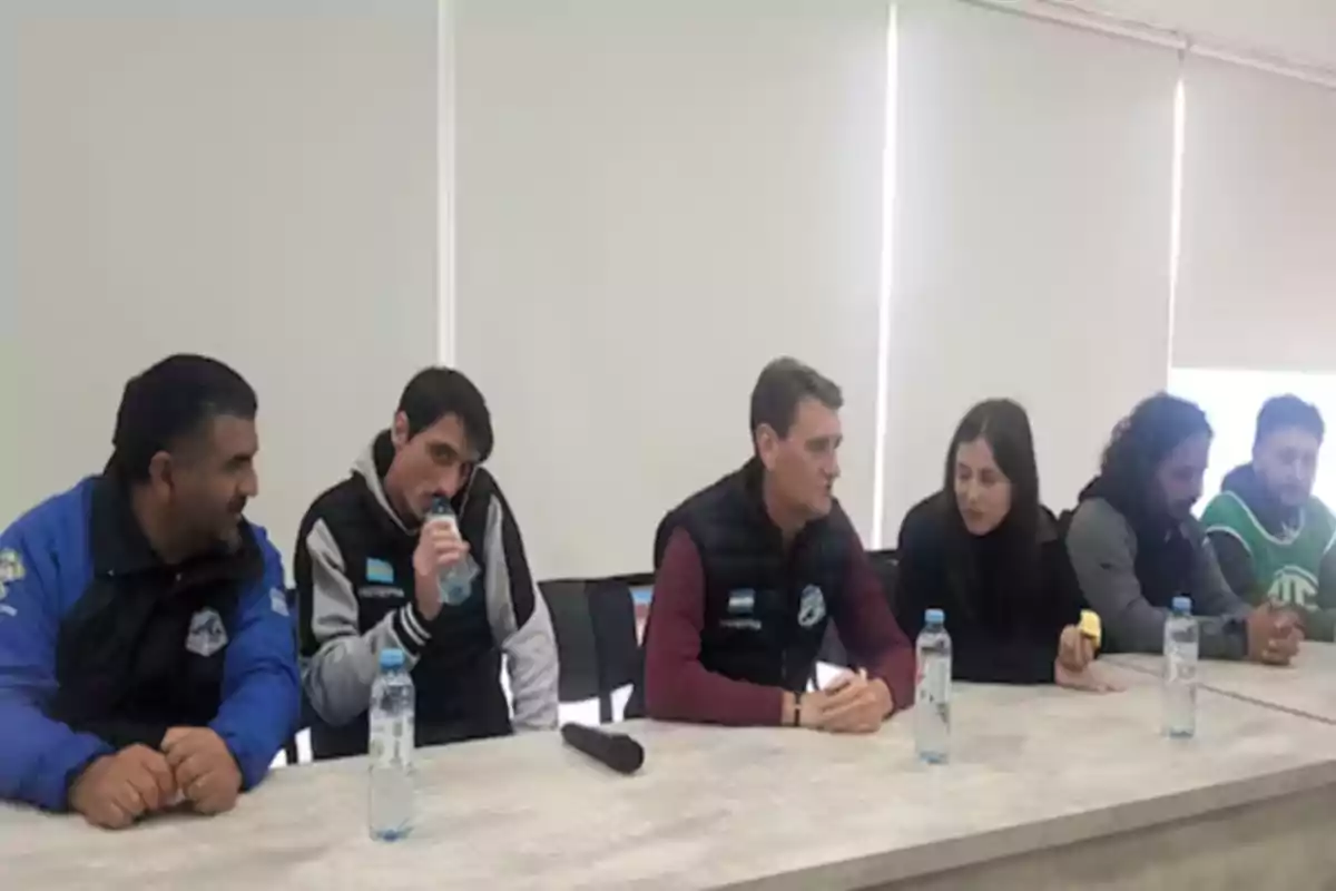 Seis personas sentadas en una mesa durante una reunión, con botellas de agua frente a ellas y una ventana con persianas al fondo
