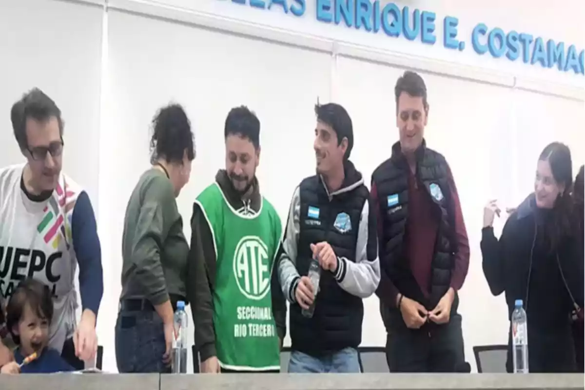 Seis personas adultas y un niño están de pie detrás de una mesa en un ambiente cerrado, algunos llevan chalecos de sindicatos y todos parecen conversar y sonreír Seis personas adultas y un niño están de pie detrás de una mesa en un ambiente cerrado, algunos llevan chalecos de sindicatos y todos parecen conversar y sonreír