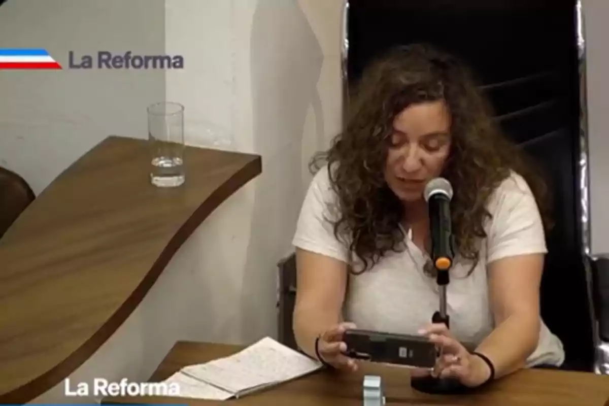 Mujer de cabello rizado sentada frente a un micrófono leyendo desde su celular en una mesa de madera con un vaso de agua y una libreta Mujer de cabello rizado sentada frente a un micrófono leyendo desde su celular en una mesa de madera con un vaso de agua y una libreta