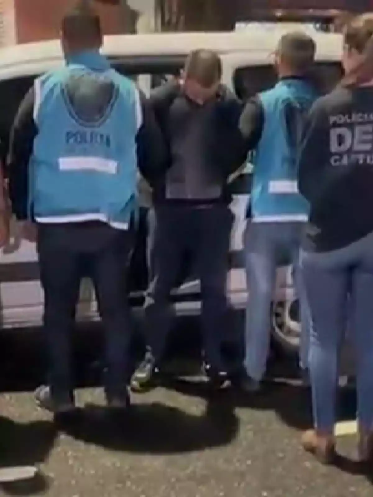Several people wearing police vests surround a handcuffed man next to a white vehicle on the street. Several people wearing police vests surround a handcuffed man next to a white vehicle on the street.