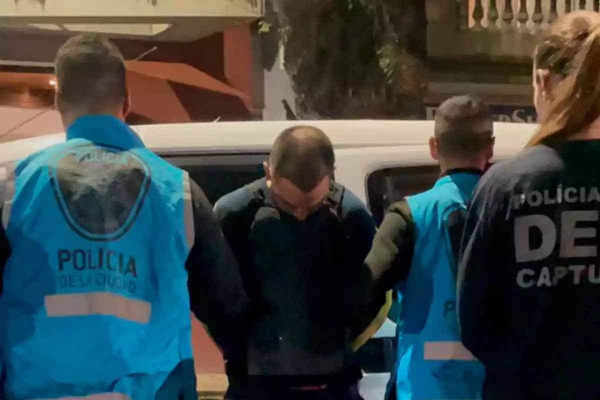 A handcuffed man is escorted by several police officers wearing blue vests in front of a white vehicle on an urban street at night.
