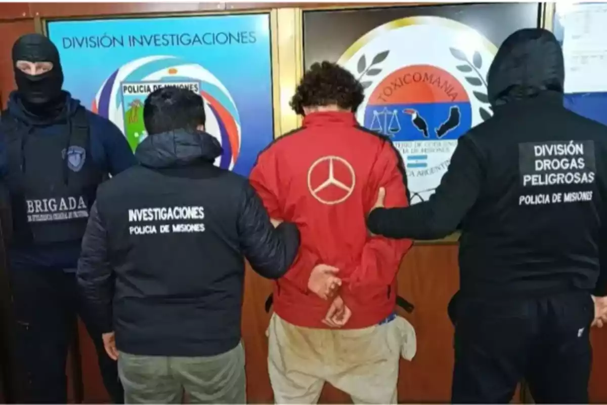 Four people with their backs turned, three of them wearing Misiones police vests and one person in the center in a red jacket, handcuffed, in front of a background with police logos. Four people with their backs turned, three of them wearing Misiones police vests and one person in the center in a red jacket, handcuffed, in front of a background with police logos.