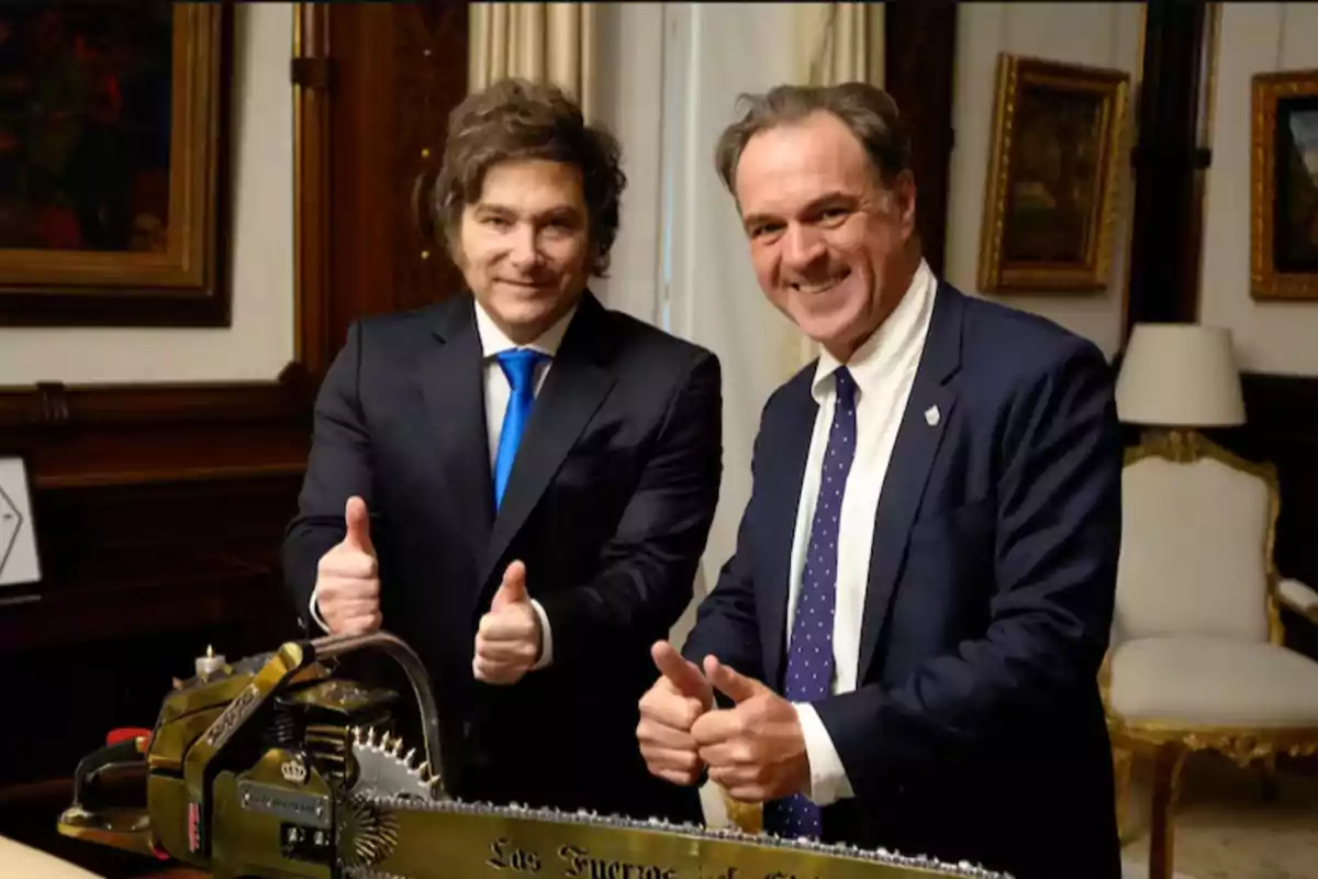 Two men in suits smile and pose with thumbs up next to a chainsaw in a stylish office.