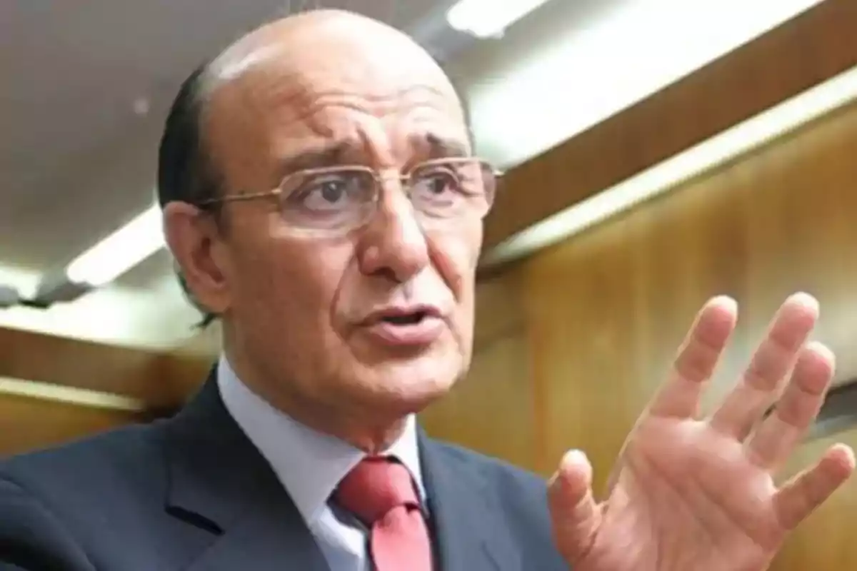 Hombre calvo con gafas y traje oscuro hablando y gesticulando con la mano en un entorno interior iluminado