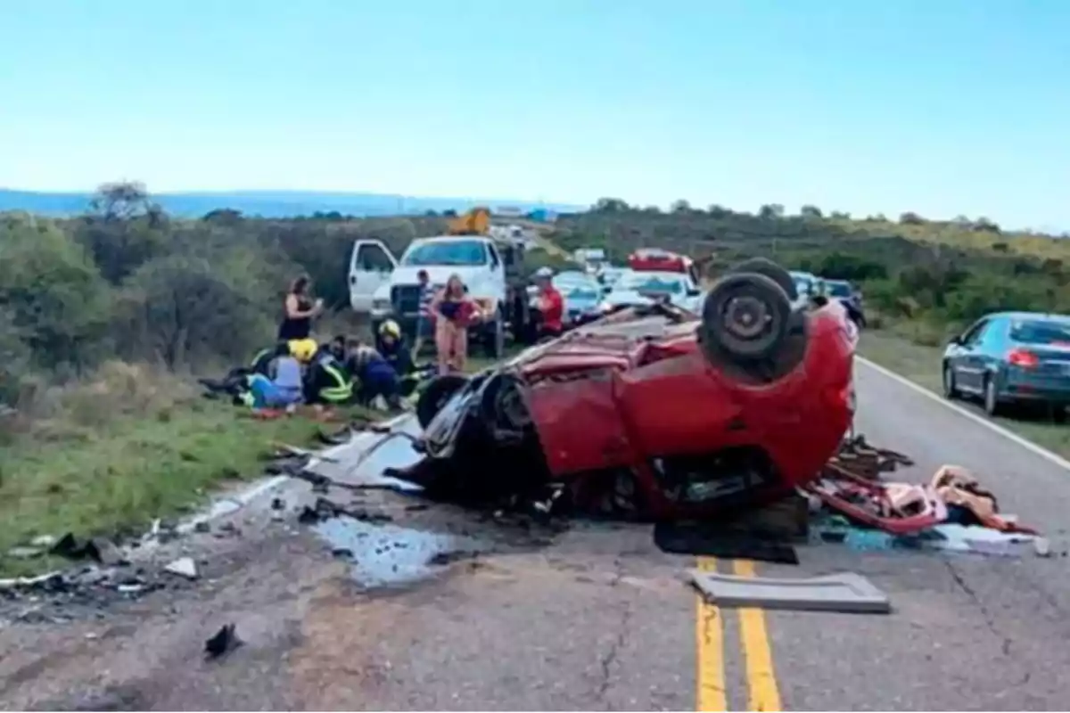 Auto rojo volcado en medio de una carretera con varios rescatistas y personas alrededor atendiendo la emergencia y otros vehículos detenidos al fondo Auto rojo volcado en medio de una carretera con varios rescatistas y personas alrededor atendiendo la emergencia y otros vehículos detenidos al fondo