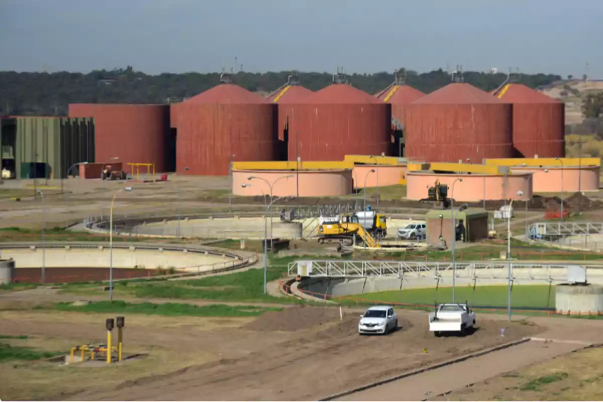 Vista de una planta de tratamiento de aguas residuales con tanques circulares grandes de color rojo y estructuras industriales rodeadas de vehículos y maquinaria Vista de una planta de tratamiento de aguas residuales con tanques circulares grandes de color rojo y estructuras industriales rodeadas de vehículos y maquinaria
