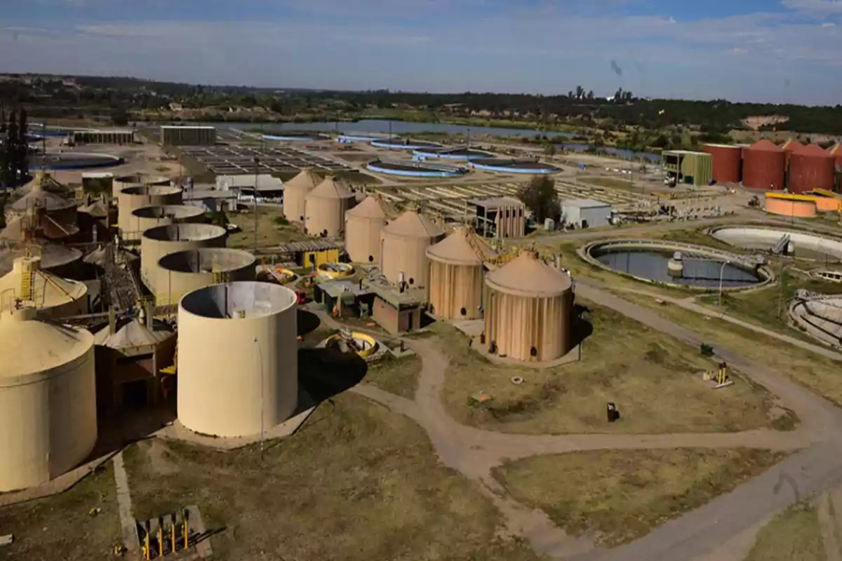 Aerial view of a wastewater treatment plant with several circular tanks and industrial structures surrounded by green areas and dirt roads