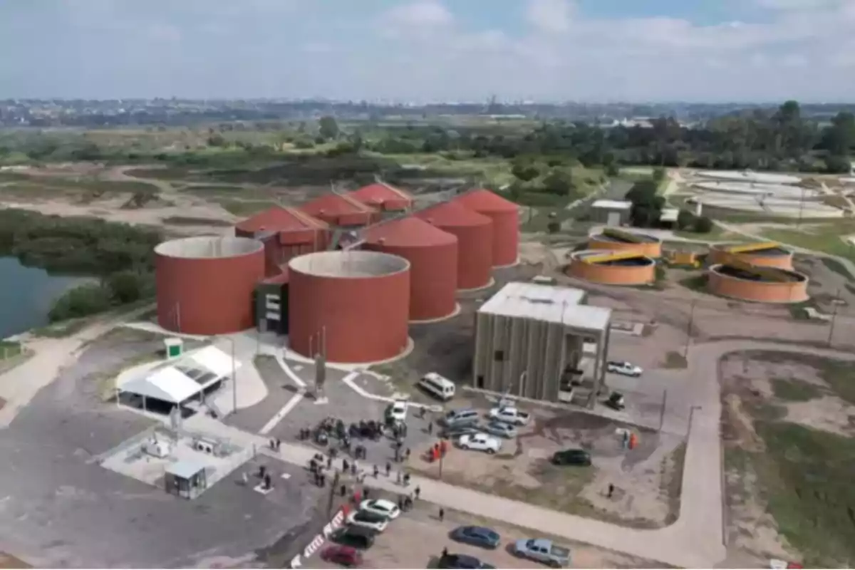 Vista aérea de una planta de tratamiento de aguas residuales con varios tanques circulares grandes de color rojo, edificios auxiliares, automóviles estacionados y personas reunidas en el área principal, rodeada de vegetación y caminos de tierra Vista aérea de una planta de tratamiento de aguas residuales con varios tanques circulares grandes de color rojo, edificios auxiliares, automóviles estacionados y personas reunidas en el área principal, rodeada de vegetación y caminos de tierra