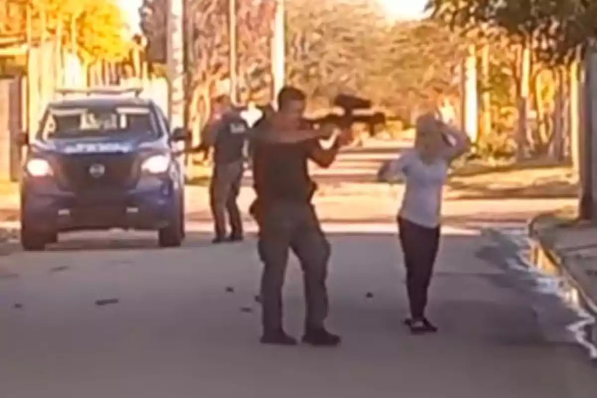 A police officer points a long gun while a woman raises her hands on a residential street and a patrol car is parked in the background.