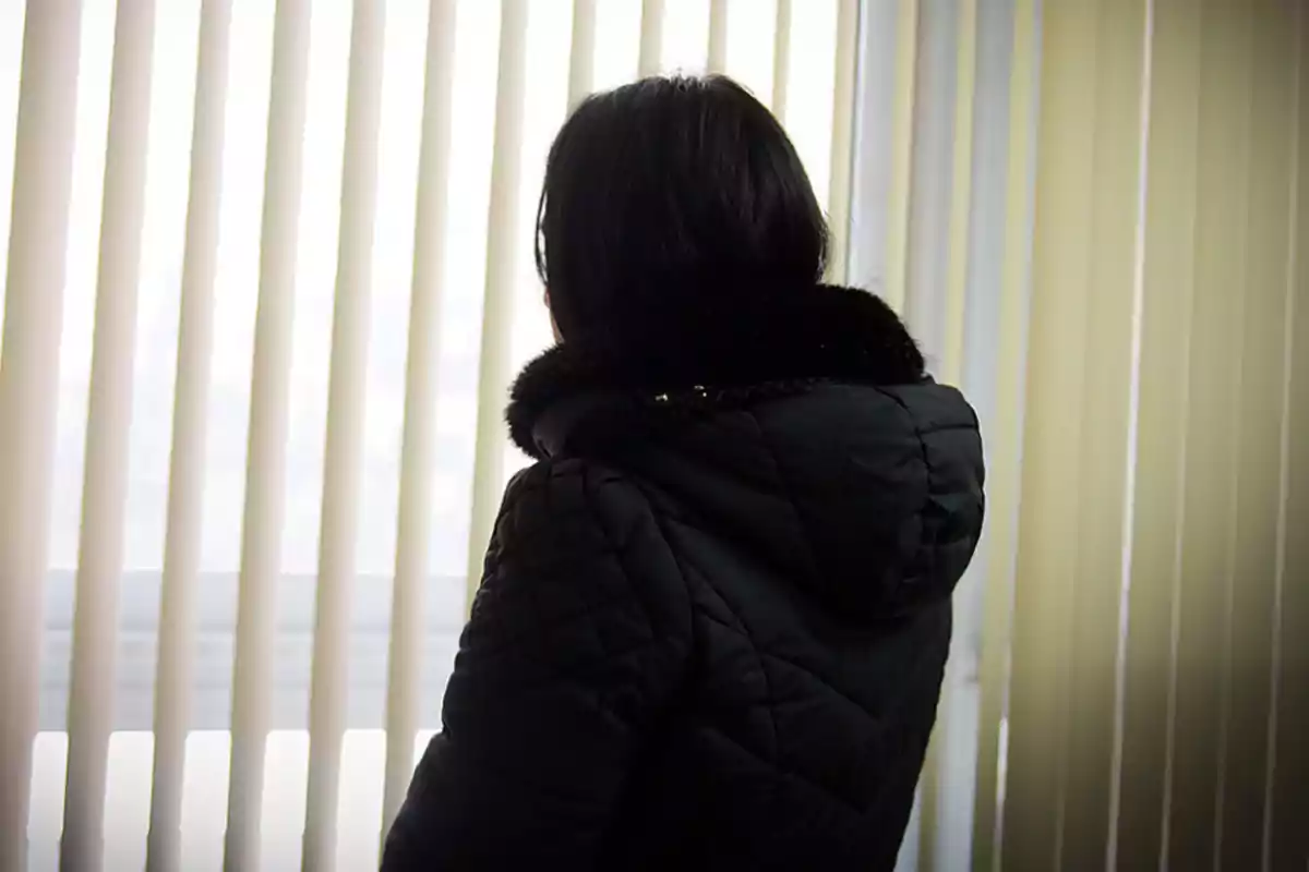 Person with their back turned wearing a dark jacket looking at a window with light vertical blinds Person with their back turned wearing a dark jacket looking at a window with light vertical blinds