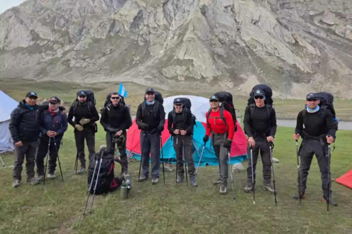 Grupo de excursionistas con mochilas y bastones de senderismo posando frente a tiendas de campaña en un paisaje montañoso Grupo de excursionistas con mochilas y bastones de senderismo posando frente a tiendas de campaña en un paisaje montañoso