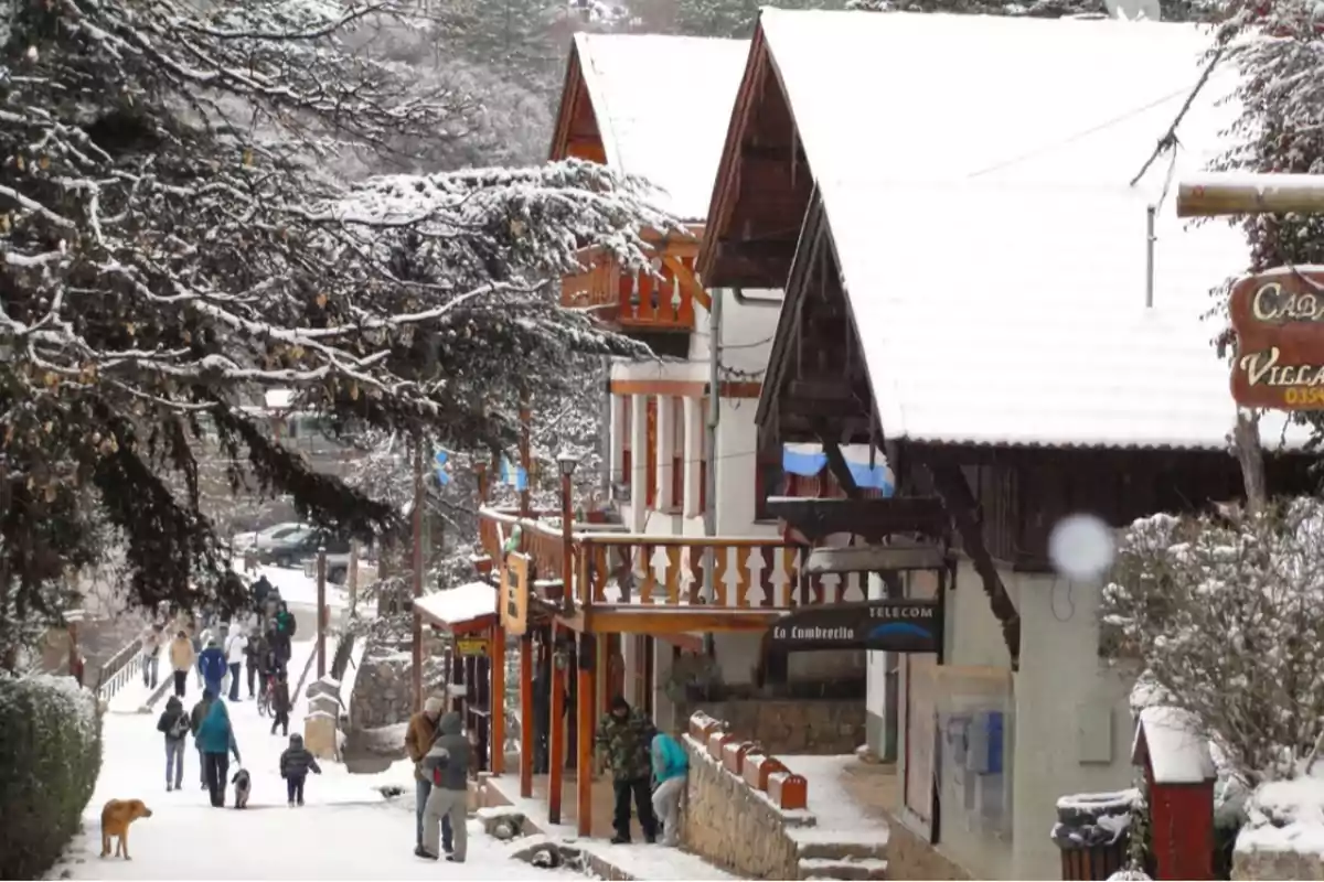Calle de un pueblo con casas de estilo alpino cubiertas de nieve, personas caminando y un perro en primer plano