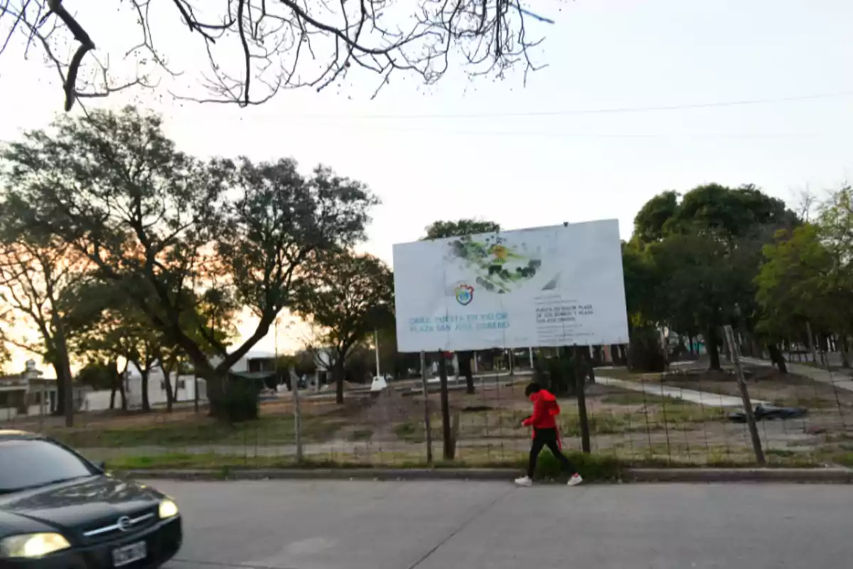 Una persona con buzo rojo camina por la vereda junto a una plaza en remodelación donde hay un cartel grande anunciando la obra, rodeada de árboles y al atardecer.
