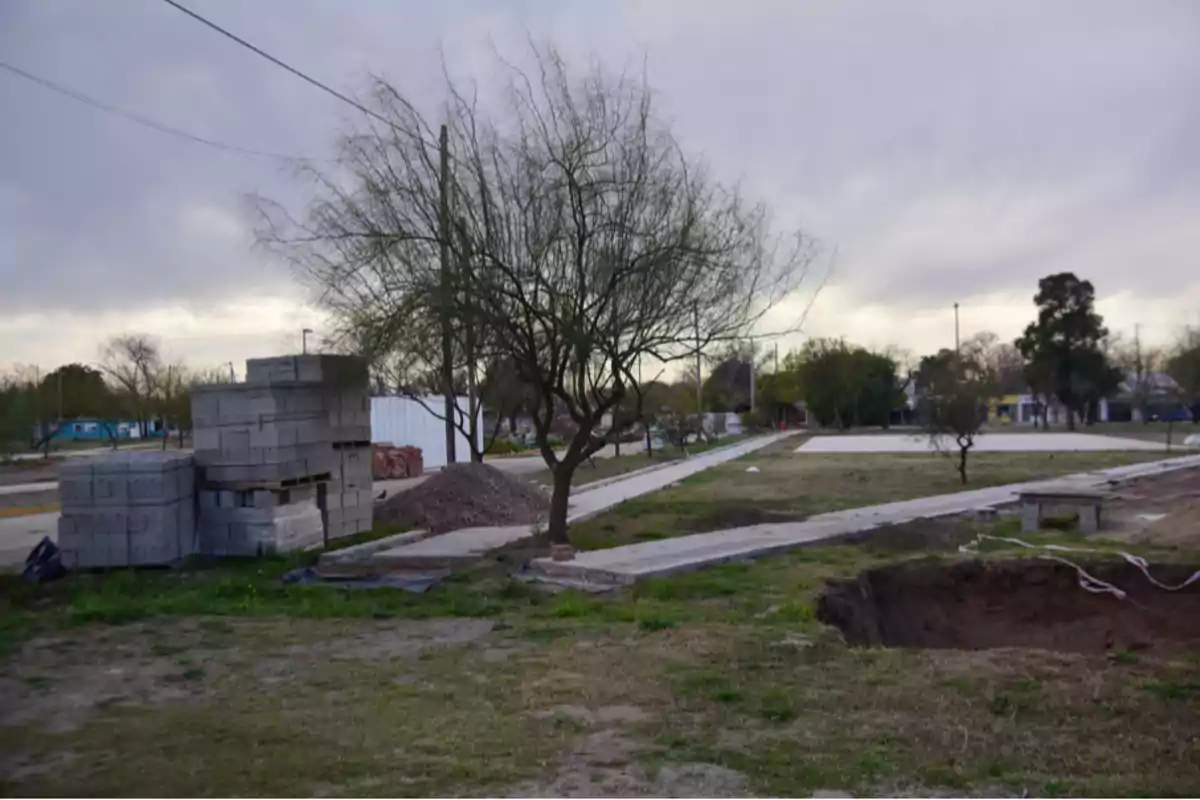 Terreno en construcción con bloques de cemento apilados, un árbol en el centro y un gran pozo en primer plano bajo un cielo nublado Terreno en construcción con bloques de cemento apilados, un árbol en el centro y un gran pozo en primer plano bajo un cielo nublado