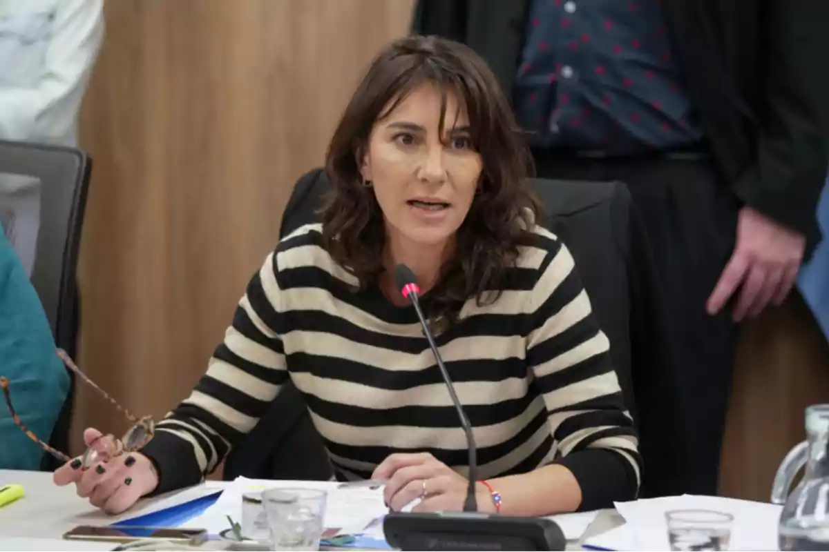 Brown-haired woman wearing a black and beige striped sweater speaking into a microphone at a meeting, holding glasses in her left hand and papers on the table