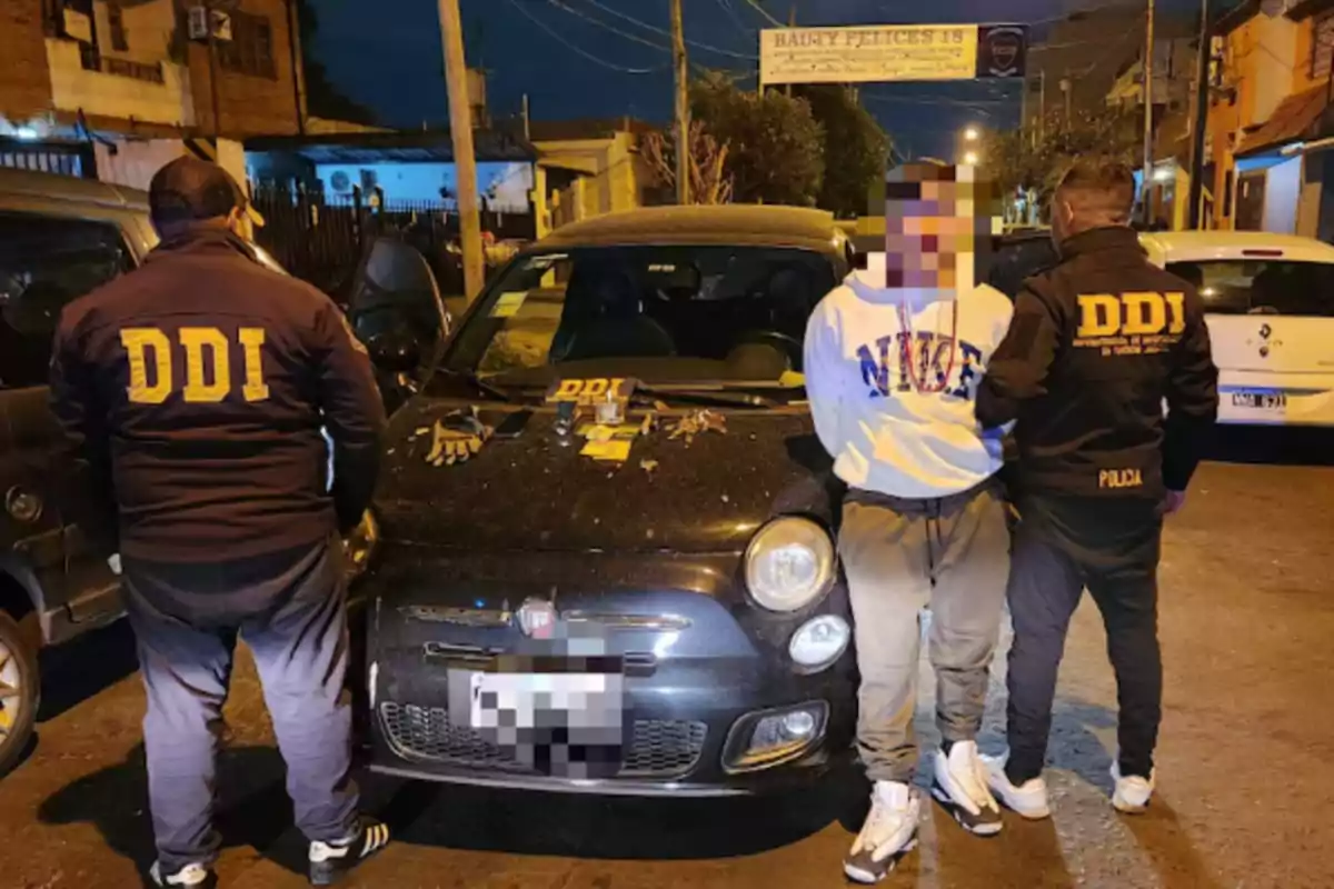 Two police officers wearing jackets that say DDI detain a person in front of a black car at night, with tools and gloves on the hood of the vehicle on an urban street. Two police officers wearing jackets that say DDI detain a person in front of a black car at night, with tools and gloves on the hood of the vehicle on an urban street.