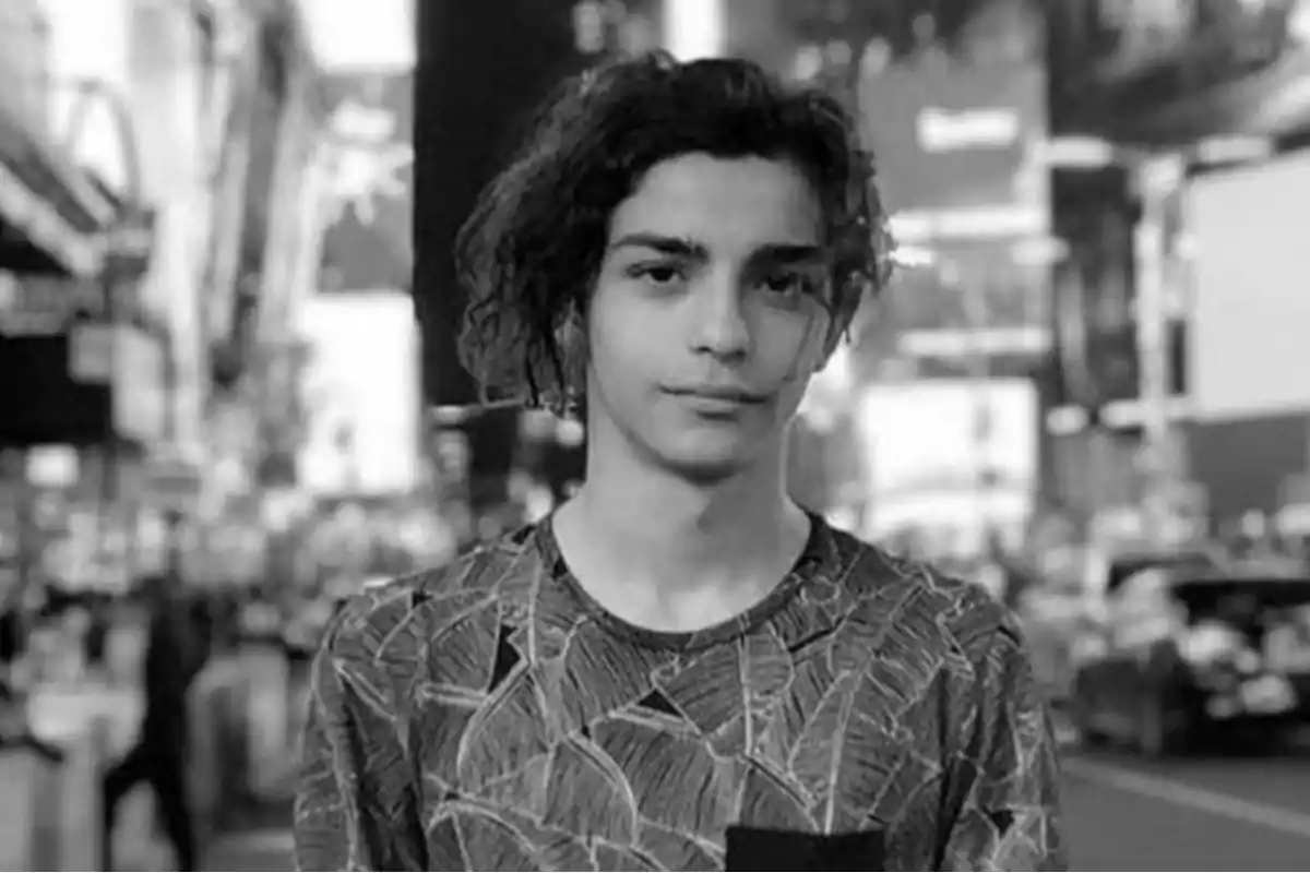 Young man with curly hair and a printed t-jersey posing in a city with a blurred black and white urban background