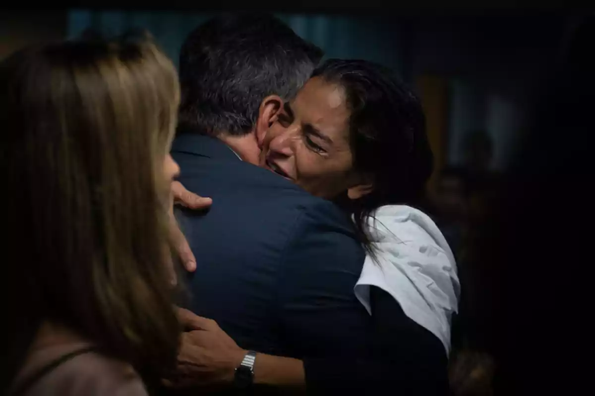 A woman hugs a man tightly and emotionally while another person watches in the foreground. A woman hugs a man tightly and emotionally while another person watches in the foreground.