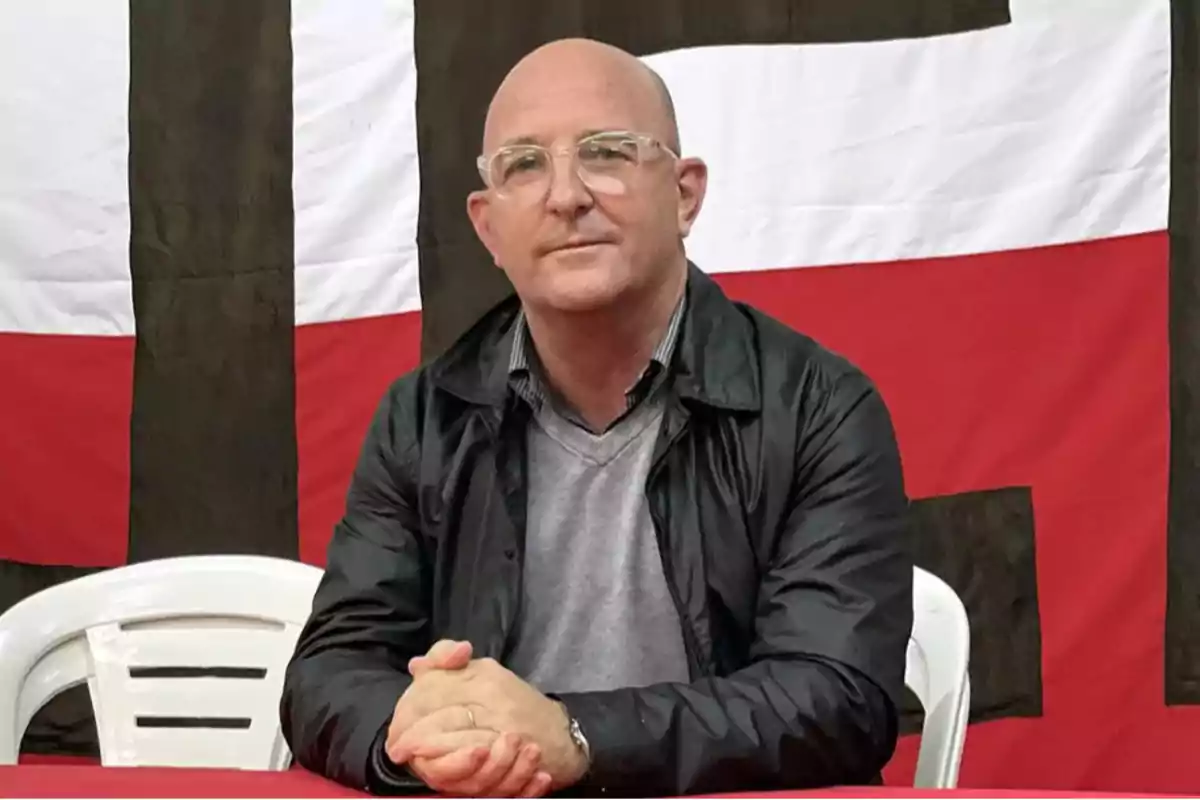 Bald man with clear glasses and a black jacket sitting in front of a red table with a flag in the background Bald man with clear glasses and a black jacket sitting in front of a red table with a flag in the background