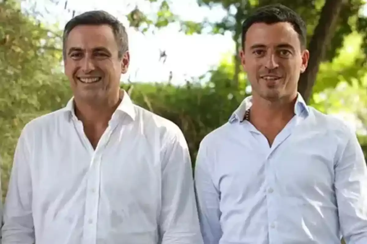 Two dark-haired men in white jerseys are standing together outdoors with trees in the background. Two dark-haired men in white jerseys are standing together outdoors with trees in the background.