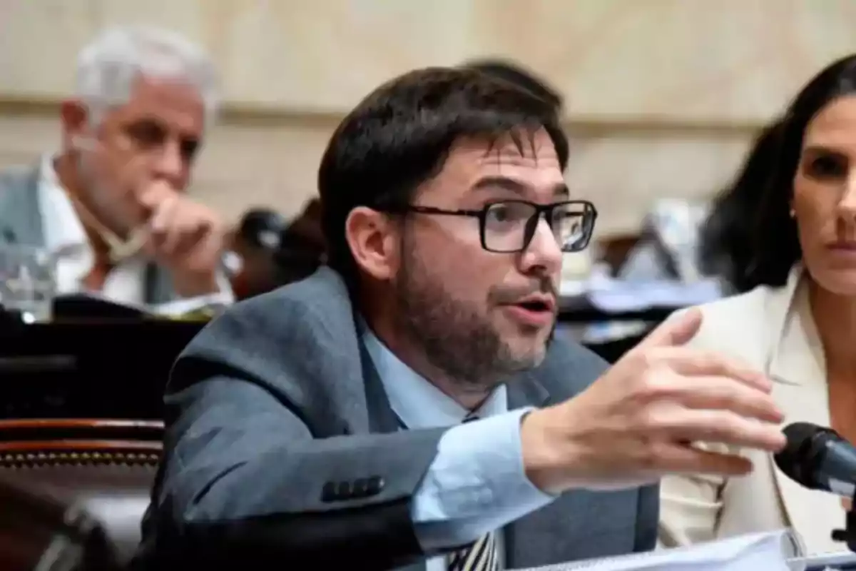 Hombre con gafas y barba hablando en una reunión formal junto a otras personas Hombre con gafas y barba hablando en una reunión formal junto a otras personas