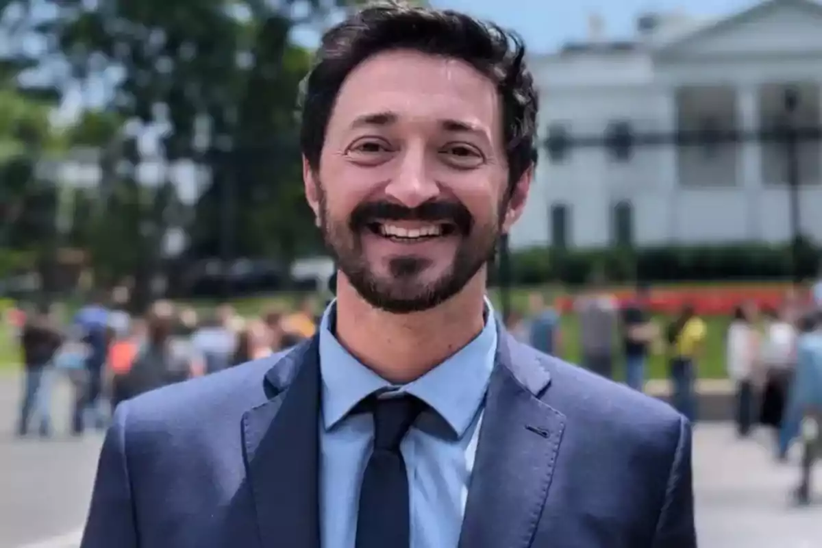 Hombre joven con barba y bigote sonriendo, vestido con traje azul y corbata, al aire libre con personas y un edificio al fondo. Hombre joven con barba y bigote sonriendo, vestido con traje azul y corbata, al aire libre con personas y un edificio al fondo.
