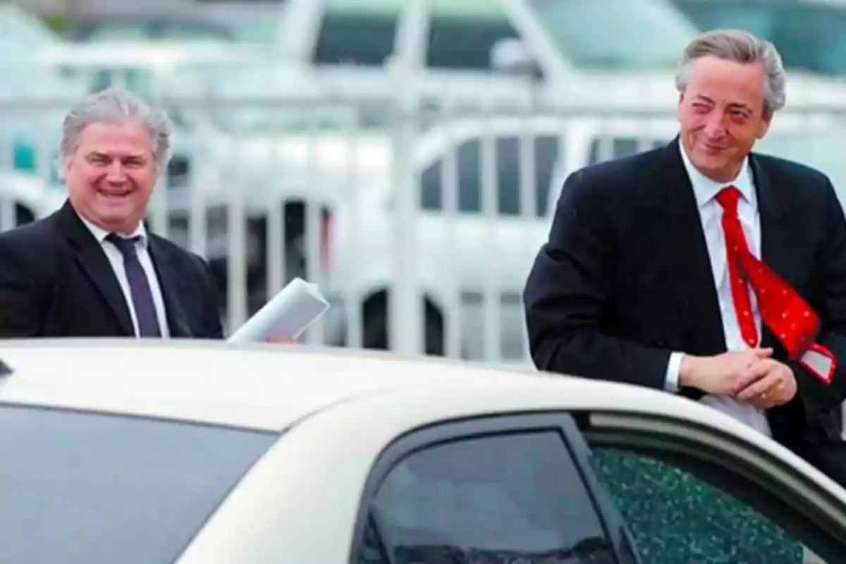 Two men in suits smile next to a white car in an urban setting with parked cars in the background.