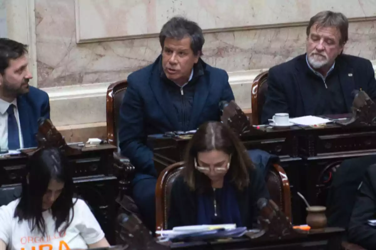 Varias personas sentadas en bancas de un recinto parlamentario, algunas conversando y otras leyendo documentos. Varias personas sentadas en bancas de un recinto parlamentario, algunas conversando y otras leyendo documentos.