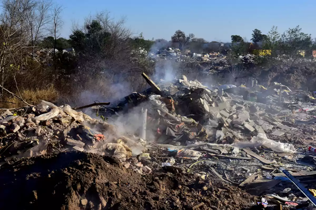 Pile of garbage and smoldering debris on an open lot surrounded by dry trees and vegetation
