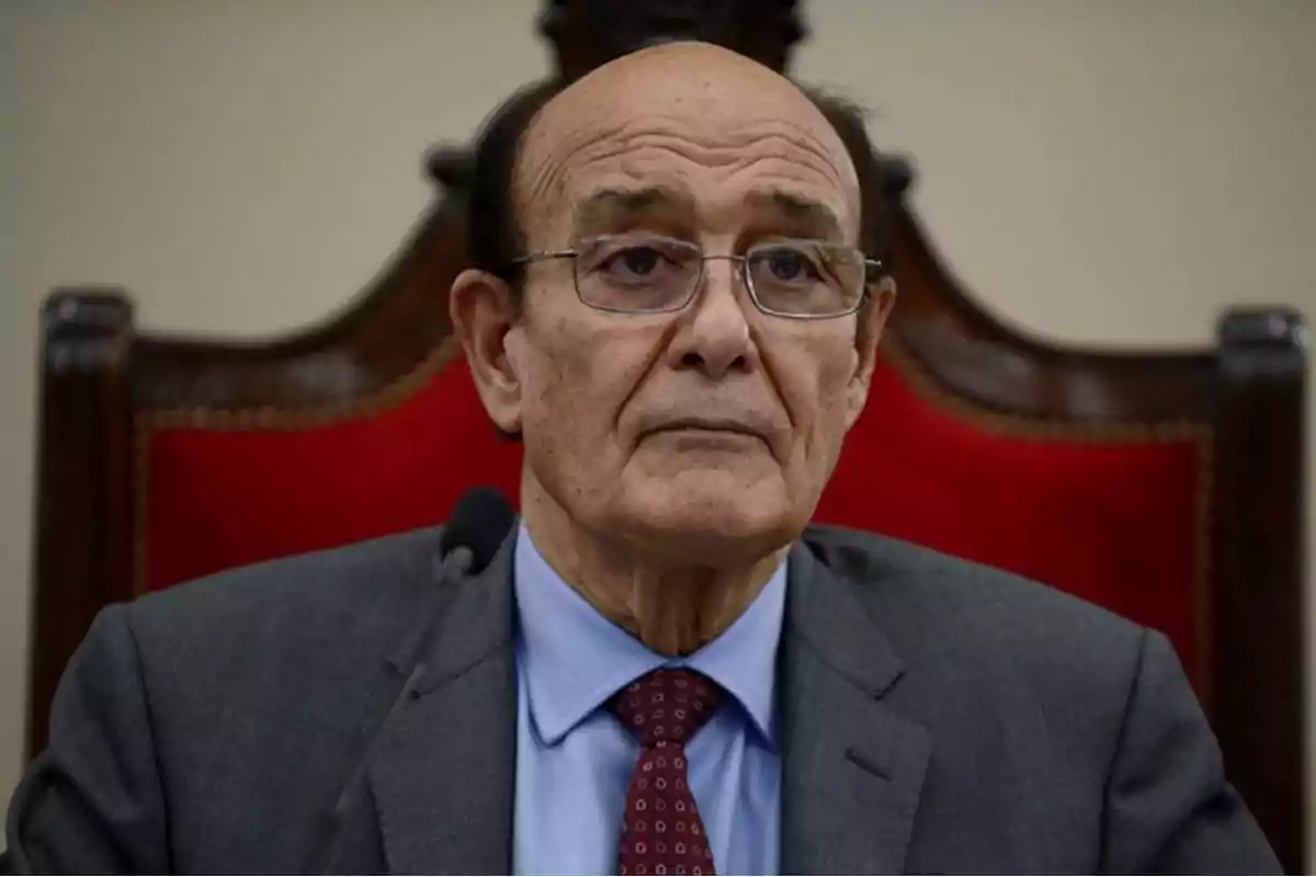 Hombre mayor con gafas y traje sentado en una silla de respaldo alto tapizada de rojo