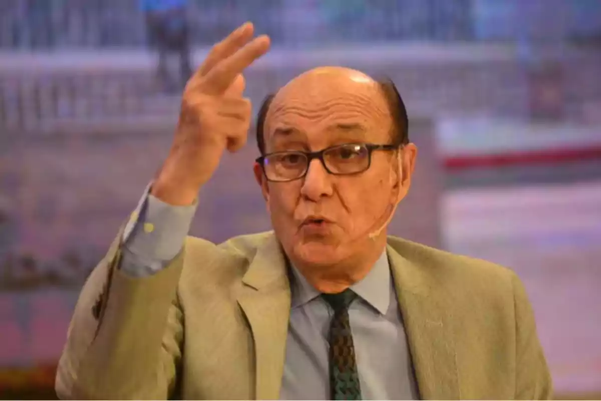 Hombre mayor con gafas y saco claro hablando en un programa de televisión mientras gesticula con la mano. Hombre mayor con gafas y saco claro hablando en un programa de televisión mientras gesticula con la mano.
