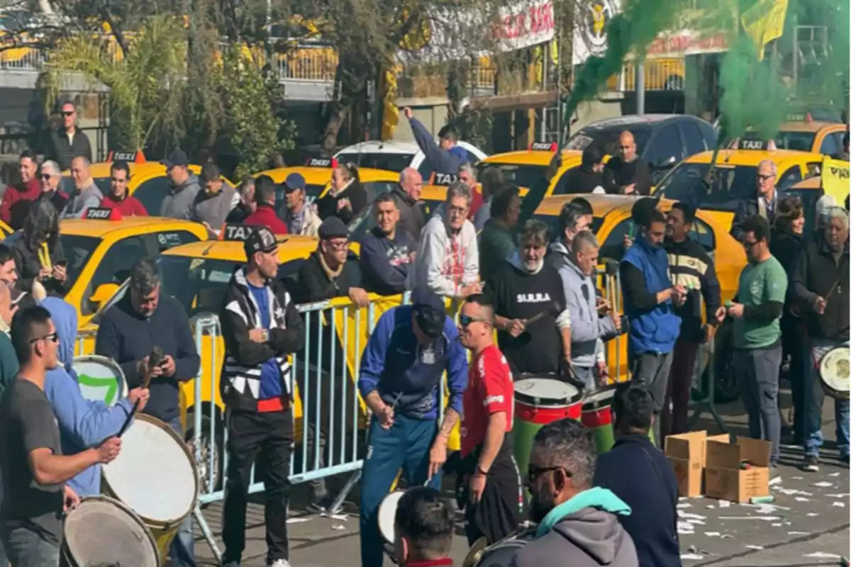 A group of people gathered in front of several yellow taxis while some play drums and green smoke can be seen in the air.