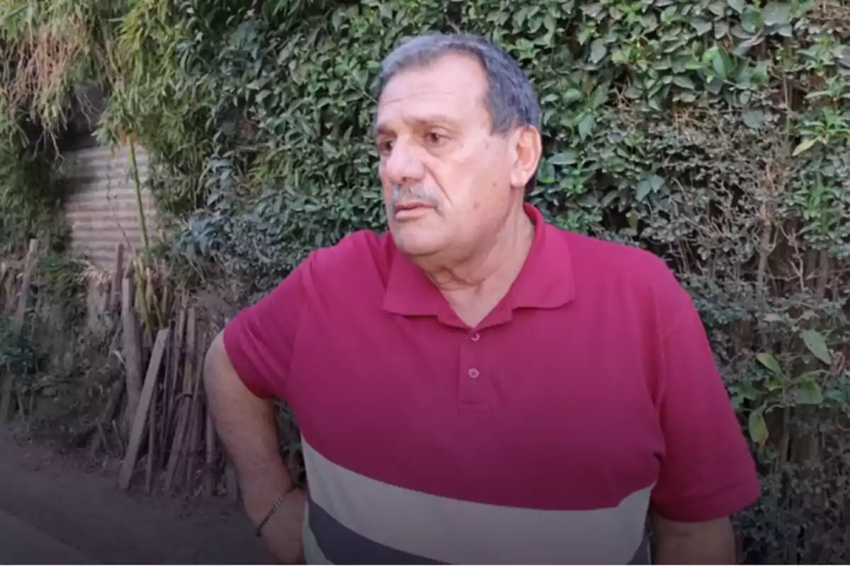 Gray-haired man with a mustache wearing a red polo jersey standing in front of a vegetation-covered fence Gray-haired man with a mustache wearing a red polo jersey standing in front of a vegetation-covered fence