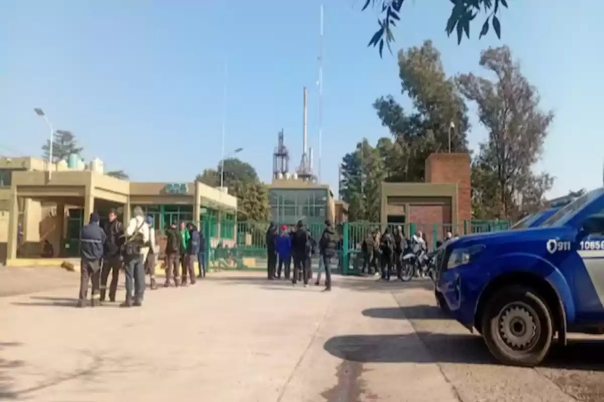 Several people gathered in front of the entrance to an industrial building with a police patrol car parked on the right.