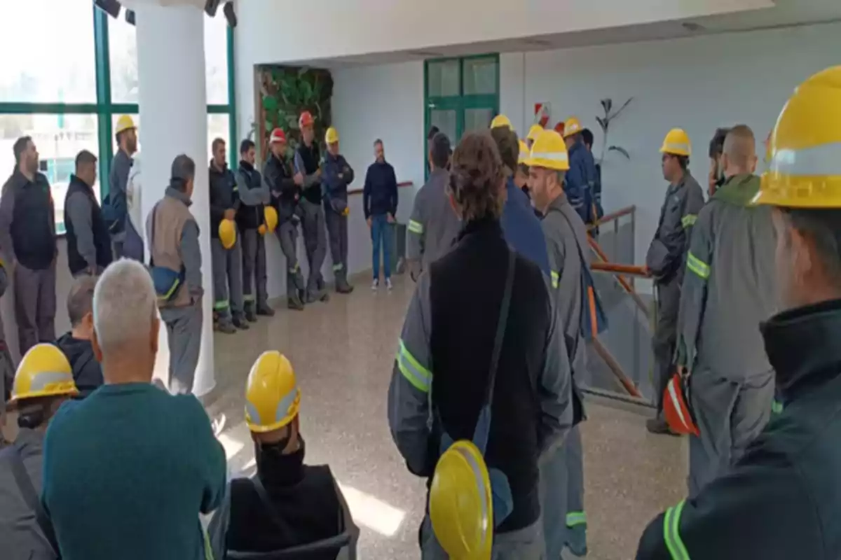 A group of workers wearing helmets and safety uniforms is gathered inside a building, some standing and others sitting, while they listen to a person who appears to be speaking in front of them. A group of workers wearing helmets and safety uniforms is gathered inside a building, some standing and others sitting, while they listen to a person who appears to be speaking in front of them.