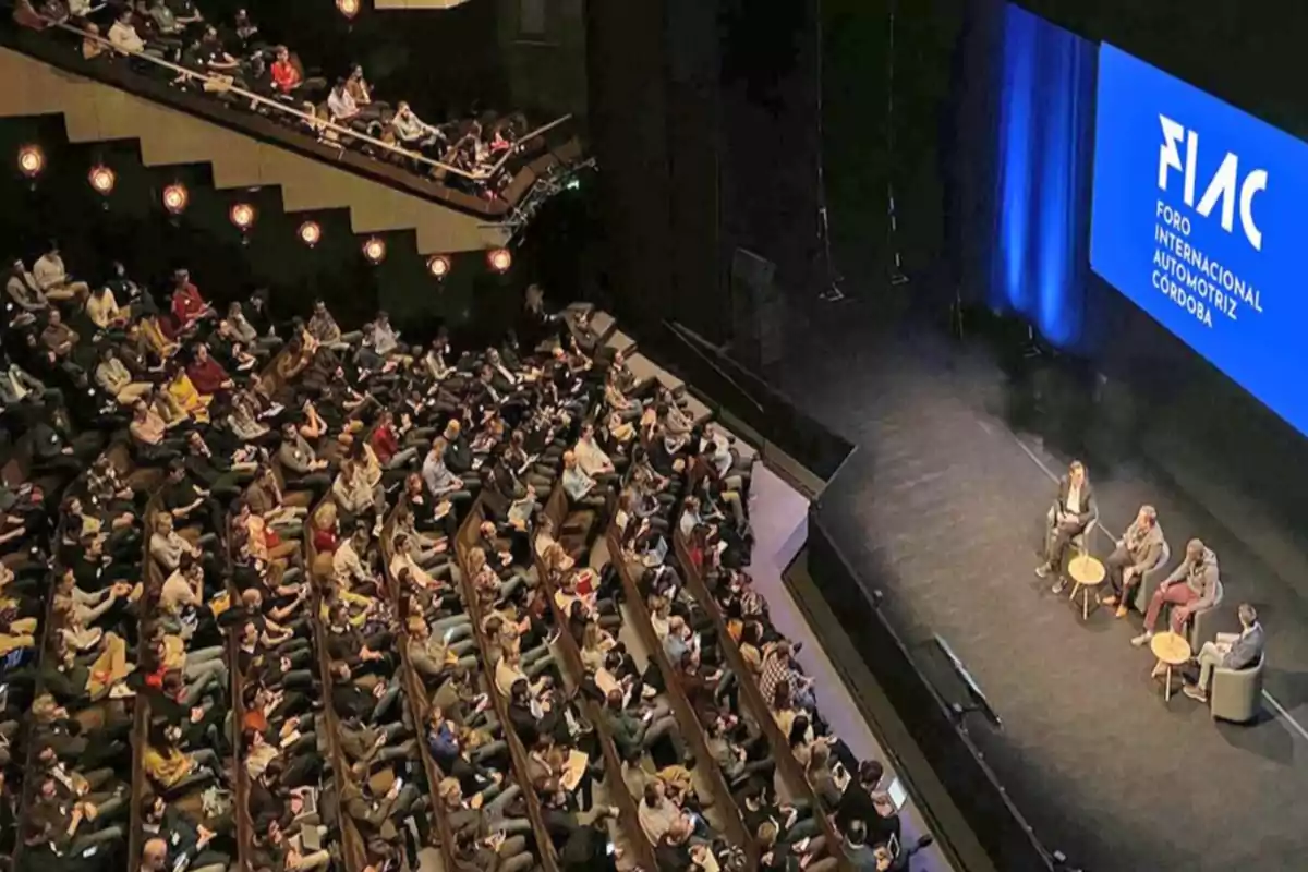 Auditorio lleno de personas asistiendo a una conferencia con cuatro panelistas en el escenario y una pantalla azul que muestra el nombre del evento FIAC Foro Internacional Automotriz Córdoba