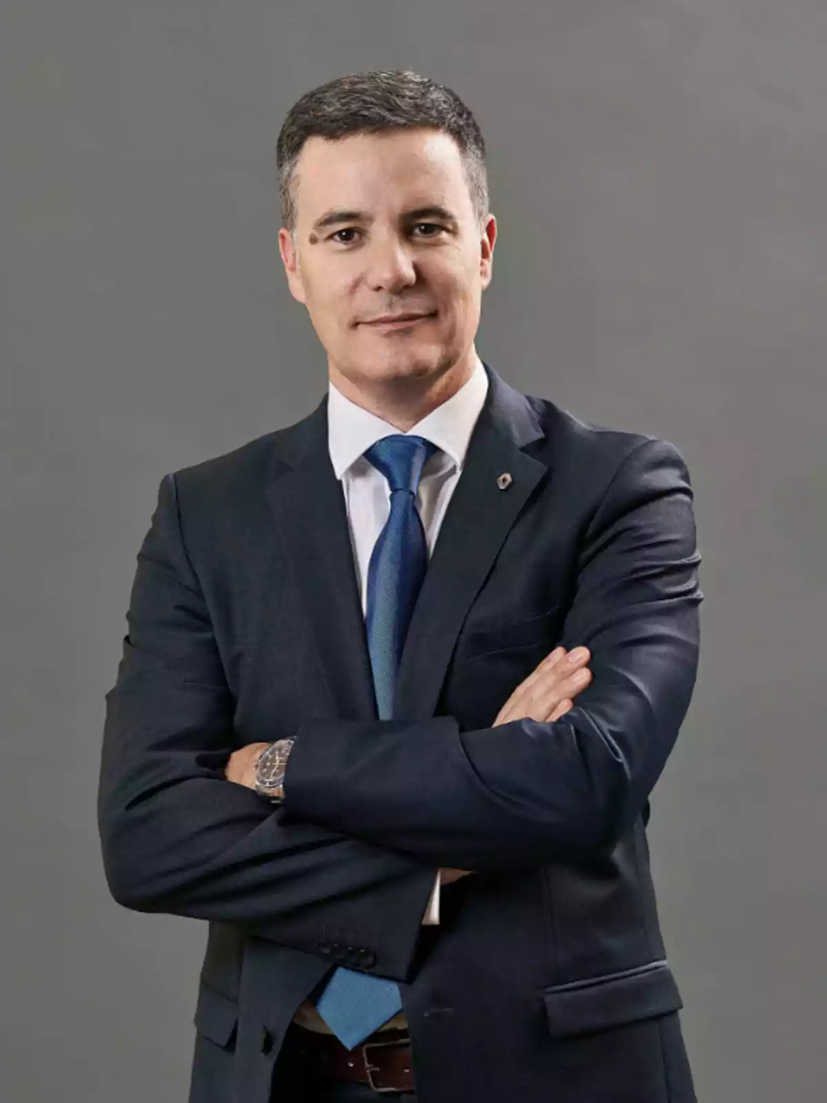 Hombre de cabello corto y canoso vistiendo traje azul oscuro con corbata azul y camisa blanca, posando con los brazos cruzados sobre un fondo gris Hombre de cabello corto y canoso vistiendo traje azul oscuro con corbata azul y camisa blanca, posando con los brazos cruzados sobre un fondo gris