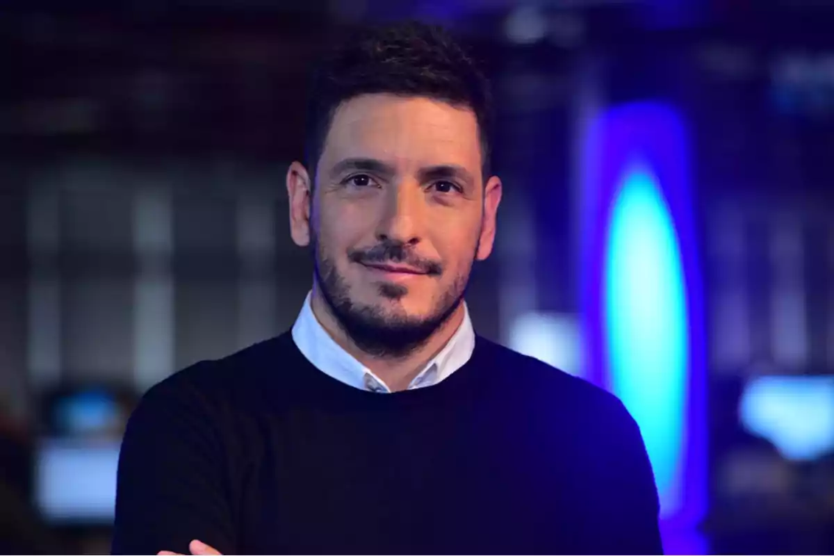 Short-haired, bearded man wearing a dark sweater and white jersey posing in an office environment with blue lights in the background